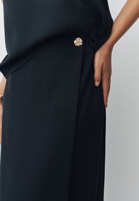 Indumento nero con tessuto liscio; presenta un accento floreale dorato vicino alla vita, con un design plissettato e una mano appoggiata sul fianco.