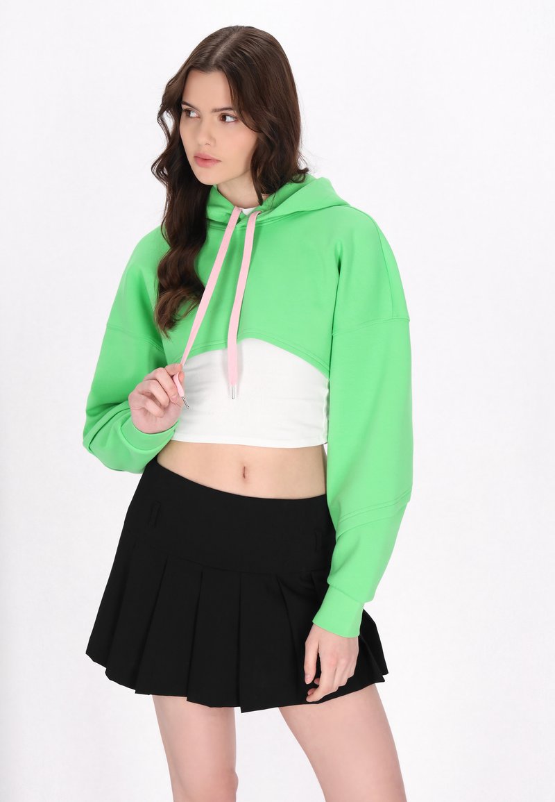 Felpa verde corta con coulisse rosa, dotata di maniche lunghe. Abbinata a una mini gonna plissè nera. Sfondo bianco.