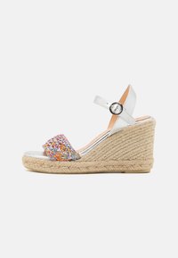 Sandale compensée blanche avec une bande textile tressée multicolore. Dote d'une bride cheville en argent et d'une semelle intermédiaire en jute de style espadrille.