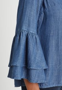 Blusa in denim blu con maniche a balze a strati, texture leggera e taglio casual. Il materiale appare leggero e traspirante.