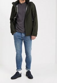 Vesitiivis oliivinvihreä huppari, harmaa t-paita, klassiset siniset farkut ja mustat lenkkarit valkoisilla yksityiskohdilla ja käännetyillä lahkeilla.