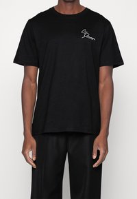 Černé bavlněné tričko s kulatým výstřihem a krátkými rukávy. Obsahuje bílé ručně psané texty na levé části hrudi.