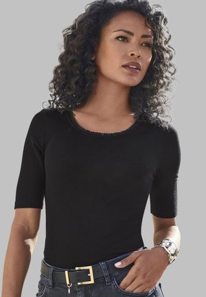 Schwarzes, geripptes Kurzarmoberteil mit einem gewellten Ausschnitt, kombiniert mit dunklen Jeans und einem schwarz-goldenen Gürtel. Trägt ein silbernes Armband.