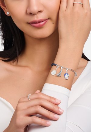 Silbernes Charm-Armband mit blauen Perlen, mehreren dekorativen Charms und einer strukturierten Kette. Am Handgelenk mit einem schlichten weißen Ärmel getragen.
