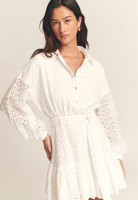 Bílé košilové šaty s vyšívaným vzorem, zapínáním na knoflíky vpředu, staženým pasem a rozšířeným lemem. Mají balónové rukávy a odnímatelný pásek.