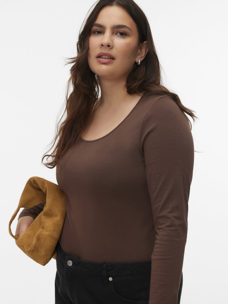 Frau mit langen braunen Haaren trägt ein enganliegendes braunes Langarmoberteil und schwarze Hosen, hält eine hellbraune Wildledertasche vor weißem Hintergrund.