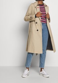 Beige trenchcoat met een kraag, dubbelgebreid ontwerp en donkere knopen, gelaagd over een rood-wit gestreept shirt en blauwe jeans.
