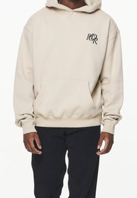 Sudadera con capucha beige, con un bolsillo frontal, puños y dobladillo acanalados, que presenta un logotipo negro en la parte superior izquierda. Textura suave, ajuste casual.