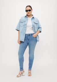 Light blue denim oversized shirt with two chest pockets, paired with fitted light blue jeans and white strappy heeled sandals.