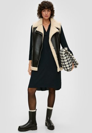 Veste en faux cuir noir avec doublure en shearling crème, portée sur une robe sombre. Le mannequin tient un sac à motif pied-de-poule. Bottines noires.