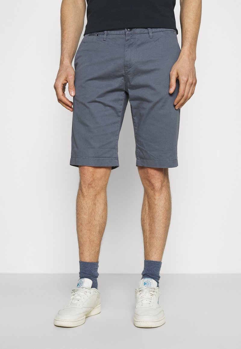 Homme portant un short gris au genou, des chaussettes foncées, des baskets blanches et un t-shirt noir, debout devant un fond blanc uni.