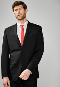 Zwarte colbert met notch-lapels, twee knopen en zijzakken; gecombineerd met een wit overhemd en een felrode stropdas, wat zorgt voor een formele uitstraling.