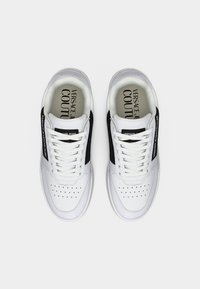 Ζευγάρι λευκών και μαύρων αθλητικών παπουτσιών Versace Jeans Couture με διατρημένες μύτες και επώνυμα πέλματα, παρουσιασμένα από πάνω σε γκρίζο φόντο.