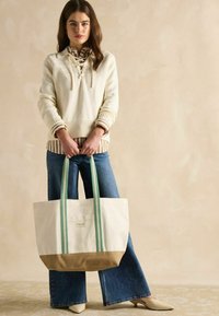 Borsa tote in tela con manici a righe verdi, fondo beige e etichetta del marchio visibile. Il modello indossa un maglione crema e jeans blu a zampa.