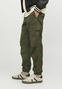 Pantalones cargo de tela verde oscuro, con múltiples bolsillos grandes, puños elásticos y un corte relajado. Llevados con zapatillas beige.
