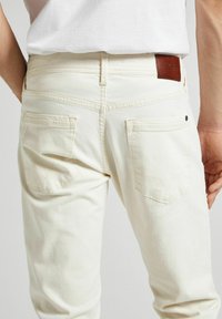 Les jeans en denim blanc présentent un design classique à cinq poches avec une étiquette en cuir à la taille. La texture est lisse avec de subtils détails de couture.