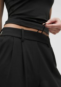 Pantaloni neri con vita elastica e pieghe, caratterizzati da una texture liscia e cordoncini regolabili, indossati sotto un top senza maniche.