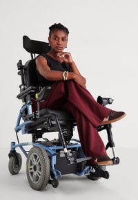 Ηλεκτρικό αναπηρικό αμαξίδιο με μπλε σκελετό και μεγάλες ελαστικές ρόδες. Χρήστης με μαύρη μπλούζα και μπορντό φαρδιά παντελόνια, καθισμένος άνετα.