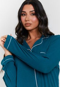 Teal button-up pajama shirt made from ribbed fabric, featuring white piping along the collar, cuffs, and pocket. Long sleeves with relaxed fit.