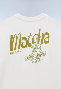Λευκό πουκάμισο με μεγάλο πράσινο κείμενο "Matcha Since 1987" και καρτουνίστικο χαρακτήρα φλυτζανιού matcha που κρατάει μια πινακίδα με τη λέξη "Πρέσβης".