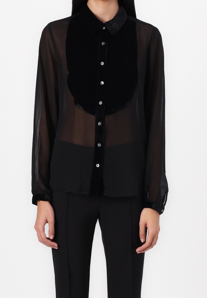 Femme portant un chemisier noir transparent à manches longues avec col en velours et patte de boutonnage, associé à un pantalon noir sur fond blanc.