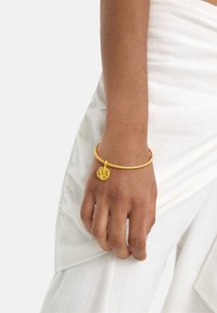 Złoty metalowy bransoletka w formie bangle z teksturowanym okrągłym charms, noszona na dłoni. Bransoletka ma prosty, okrągły kształt i gładkie wykończenie.