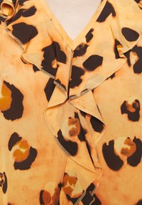 Blusa arancione con motivo leopardato, caratterizzata da scollo arricciato e maniche corte. Tessuto morbido con dettagli marroni e gialli.