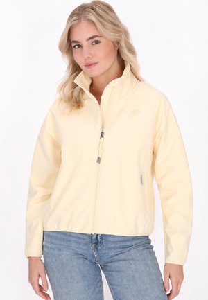 Jonge vrouw met golvend blond haar draagt een lichtgele jas met rits en lichtblauwe spijkerbroek tegen een witte achtergrond.