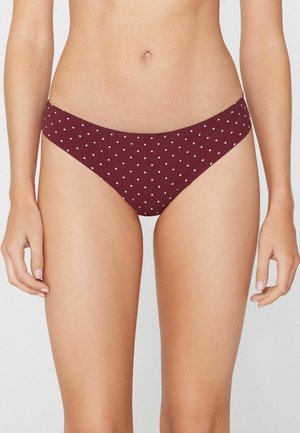 Kobieta w bordowych majtkach bikini w białe kropki, stoi z rękami luźno opuszczonymi wzdłuż boków na jednolitym tle.