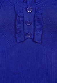 Modrý pletený top s třemi knoflíky ve tvaru růže a volánkovou texturou podél výstřihu, se savým žebrovaným vzorem látky.