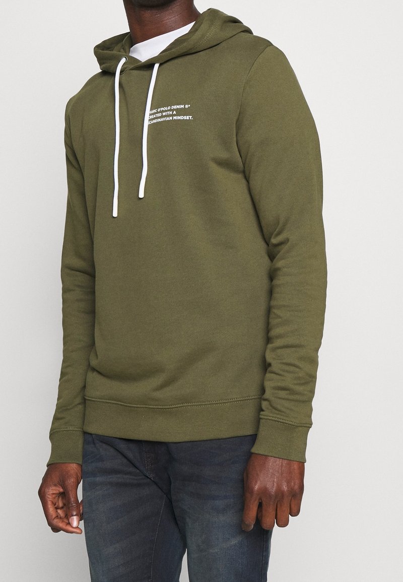 Homme portant un sweat à capuche vert olive avec des cordons blancs et un jean bleu foncé, debout sur un fond clair uni.