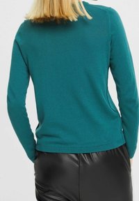 Türkiser Pullover mit langen Ärmeln und rundem Ausschnitt, aus weichem Strickstoff; kombiniert mit schwarzen Kunstlederhosen.
