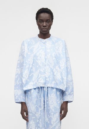 Osoba ubrana w jasno niebieski strój z białymi kwiatowymi wzorami, w tym zapinany na guziki top i pasujące spodnie, stojąca na jednolitym tle.