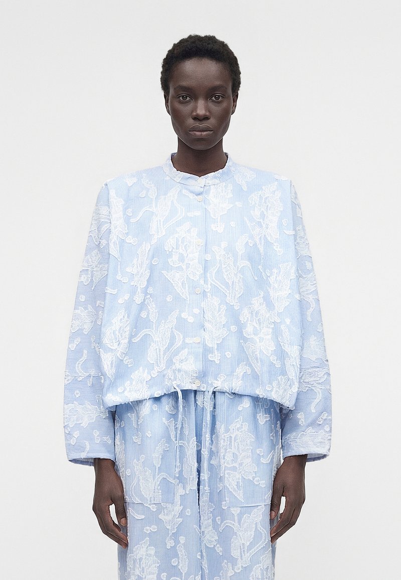 Άτομο που φοράει ανοιχτό μπλε ρούχο με λευκά λουλουδάτα σχέδια, που περιλαμβάνει ένα πουκάμισο με κουμπιά και ταιριαστά κάτω μέρη, στέκεται μπροστά σε απλό φόντο.