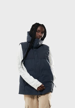 Marineblauwe puffer gilet met een hoge kraag, voorzien van twee voorzakken en een gladde, gewatteerde textuur. Draagt over een wit shirt met lange mouwen.