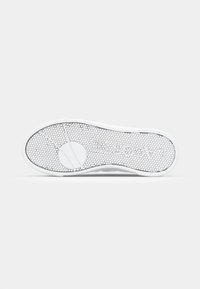 Fehér sneaker talp, amely textúrázott gumifelszínt tartalmaz, hatszögletű mintázattal és kerek logóval a közepén.