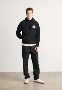 Sudadera negra con bolsillo frontal y diseño gráfico blanco, combinada con pantalones de mezclilla oscuros y zapatillas blancas con suelas de goma.