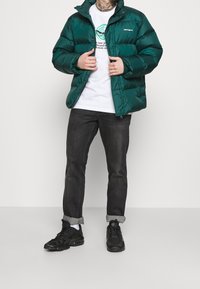 Chaqueta acolchada verde con un acabado brillante, cierre de cremallera frontal, cuello alto y logo bordado. Llevada sobre un suéter blanco y jeans oscuros.