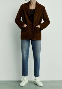 Blazer de ante marrón con solapas de muesca, dos bolsillos frontales y un ajuste relajado. Combinado con jeans rectos de tiro claro y zapatillas blancas.