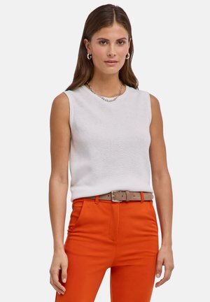 Femme aux longs cheveux bruns, portant un haut blanc sans manches, un pantalon orange, une ceinture beige et des boucles d'oreilles argentées en anneau, faisant face vers l'avant.
