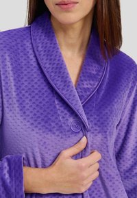 Bata de baño morada con una superficie texturizada, que presenta un cuello chal, un cierre de un solo botón y puños enrollados. Tejido suave y peludo.
