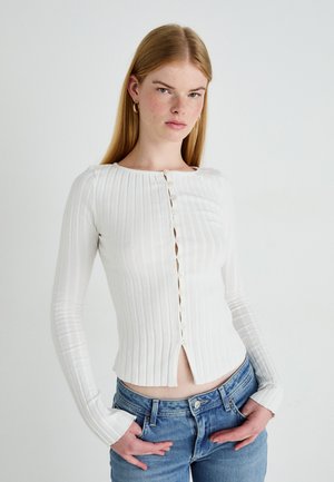 Witte geribbelde langemouwen shirt met een ronde hals en een knoopsluiting aan de voorkant. Strak ontwerp, zachte textuur en een cropped zoom, gecombineerd met blauwe jeans.
