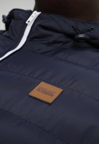 Close-up van een marineblauwe pufferjack met een bruin leren label "Urban Classics" nabij de borst en een met rits gesloten kraag.