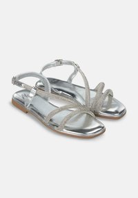 Flache Sandalen in metallischem Silber mit gekreuzten Riemen und einem strukturierten Akzent. Mit einem verstellbaren Knöchelriemen und einer leichten Sohle.