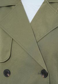 Oliivinvihreä kaksirivinen takki, jossa isot mustat napit, leveä kaulus ja sileä pinta. Saumoissa yksityiskohtaiset ompeleet.