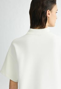 Krēmkrāsas oversize polo krekls ar teksturētu audumu, īsām piedurknēm un smalku logotipa detaļu aizmugurē pie kakla.