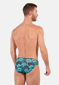 Herren-Schwimmslip mit einem floralen Muster in Türkis und Marineblau. Aus glattem Stoff gefertigt, hat er eine enganliegende Passform und moderate Abdeckung.