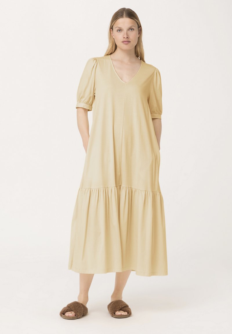 Helles beigefarbenes Maxikleid mit V-Ausschnitt, kurzen Puffärmeln und gerafftem Saum. Hergestellt aus weichem Material, mit seitlichen Taschen. Getragen mit braunen, flauschigen Hausschuhen.