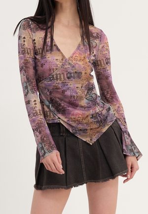 Femme portant un top cache-cœur imprimé violet, orange et rose à manches longues et une mini-jupe plissée noire avec un ourlet effiloché.