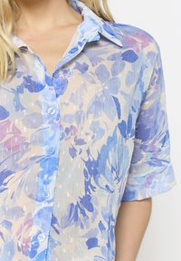 Lengvas, permatomas bliuskas su gėlių raštu mėlynos ir levandų atspalviuose, pasižymintis mygtukų užsegimu priekyje ir švelniai suktomis rankovėmis.